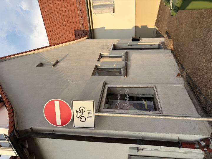 Hausecke an der Straße; rechts beginnt die Gasse, die zu beiden Eingängen führt